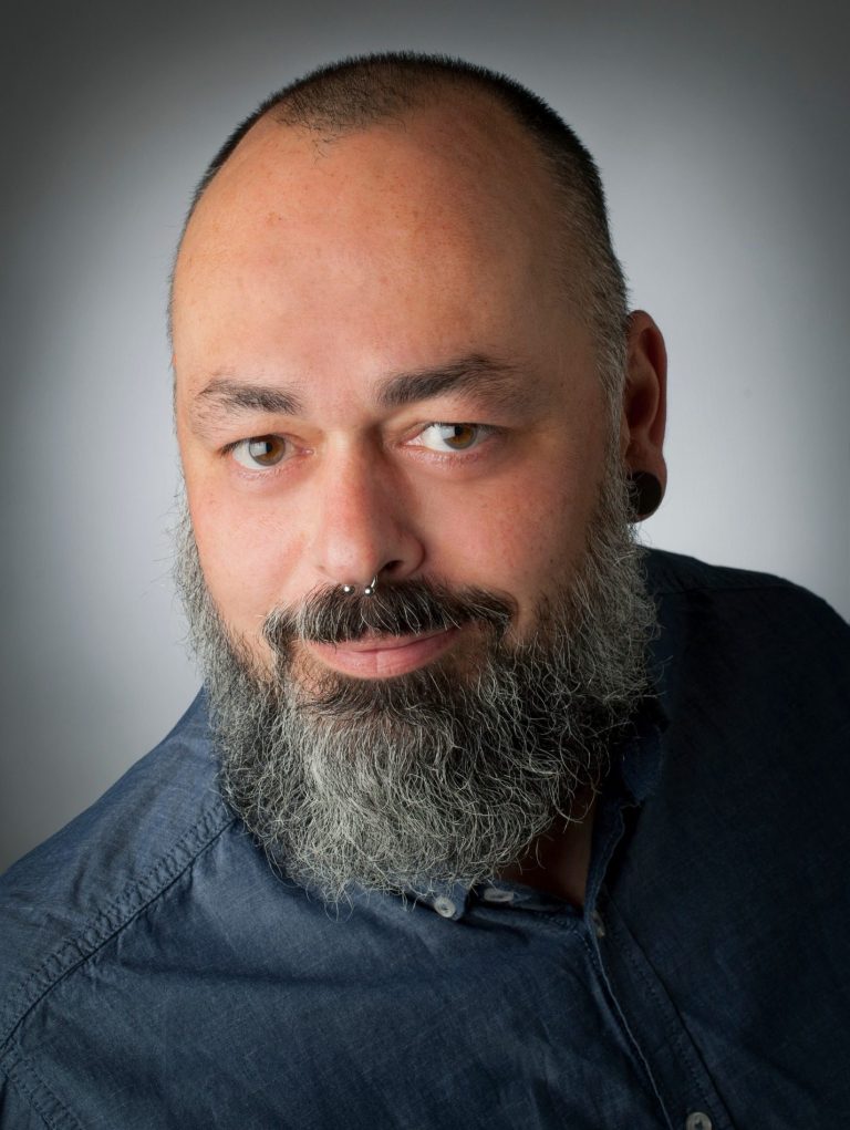 Mann mit Vollbart und kurzen Haaren, trägt ein blaues Hemd und schaut direkt in die Kamera.